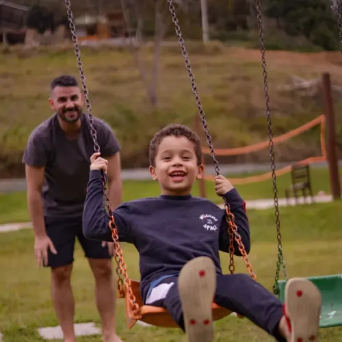 Criança sorrindo em um balanço enquanto um adulto está atrás, em um parque.