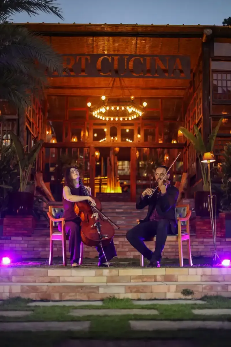 Músicos tocando violoncelo e violino em um palco iluminado ao ar livre à noite, em frente a um restaurante.