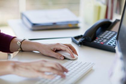 Mani di una persona che usa il mouse e la tastiera di un computer su una scrivania, telefono e raccoglitori sullo sfondo.