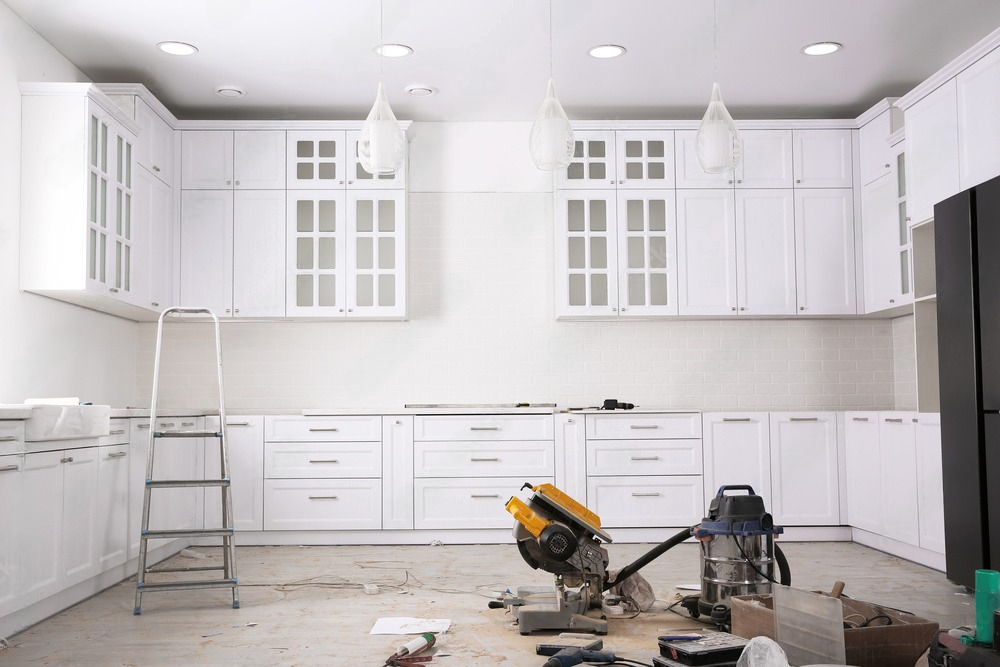 A kitchen under construction with white cabinets and a circular saw on the floor.