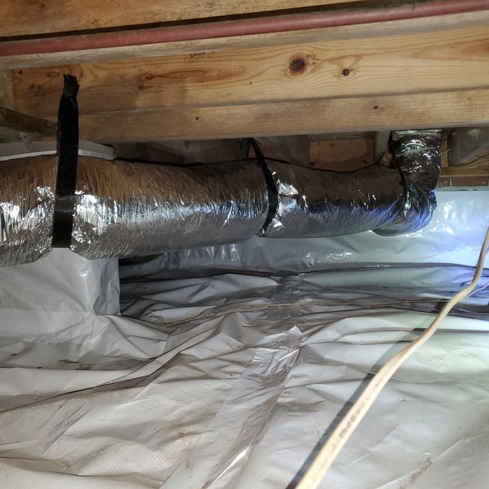 Under-house view: Ductwork wrapped with black straps, installed under wooden beams, with white plastic vapor barrier.
