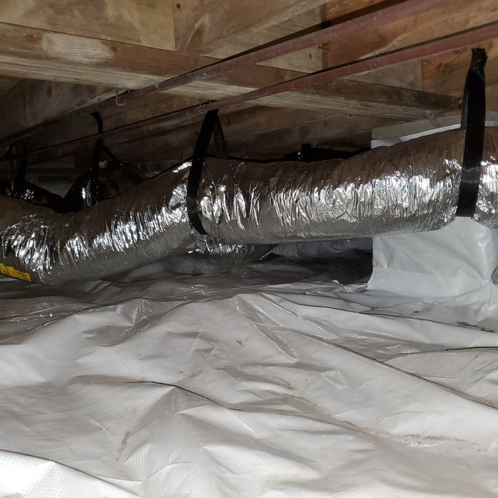 Under-house view: white vapor barrier, silver HVAC ductwork, and black straps suspended from wooden beams.