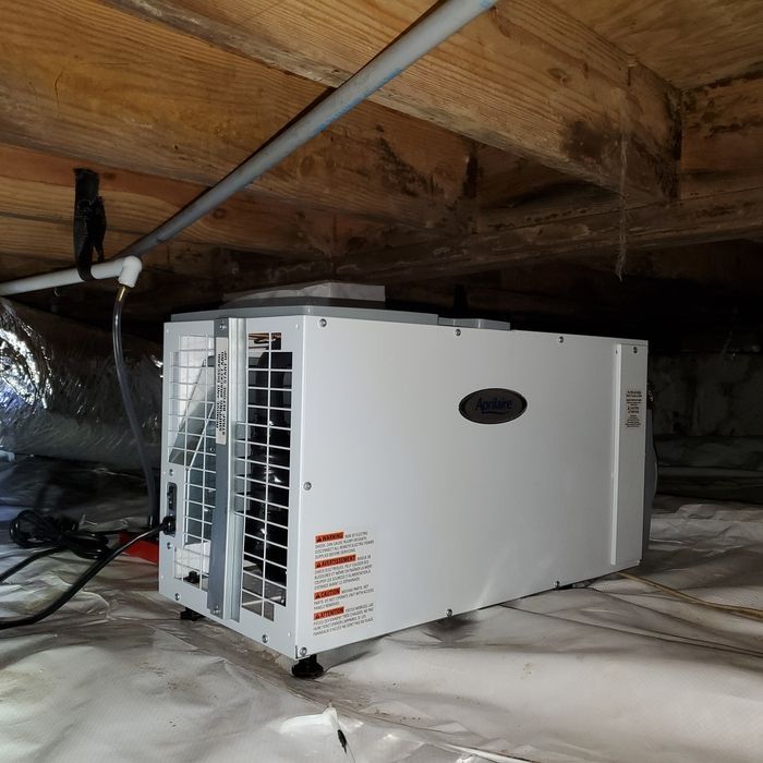 White dehumidifier in a crawl space, under wooden beams, with a white plastic cover.