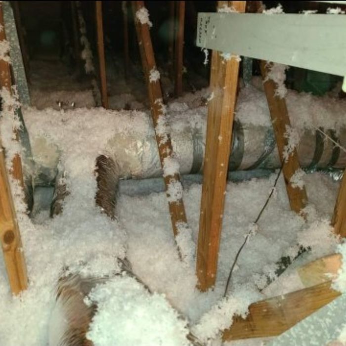 Attic insulation surrounds ductwork and wooden beams. White insulation and gray ductwork in a dark, indoor setting.
