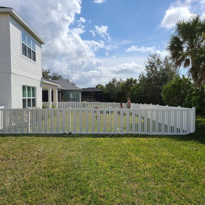 White fenced backyard with a house, blue sky, and people playing.