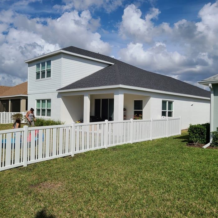 White two-story house with black roof, white fence, and green lawn under a blue sky with clouds.