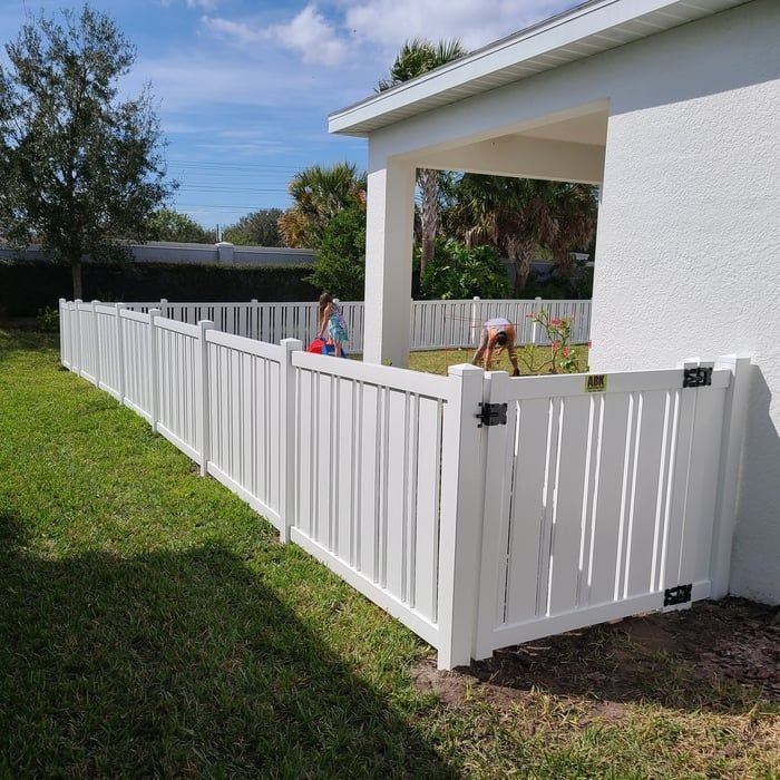 White vinyl fence surrounding a grassy backyard; two people near the house.