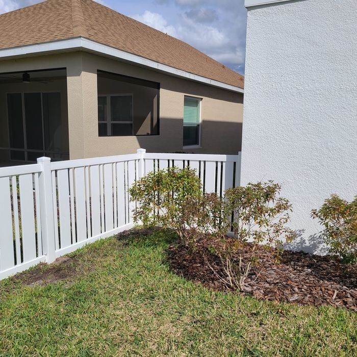 White fence surrounds a small yard with bushes, grass, and two houses in the background.