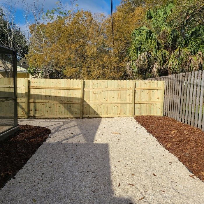 Backyard with light gravel path, wooden fence, and mulch borders. Trees in background.