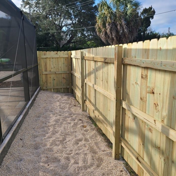 Narrow gravel pathway between a wooden fence and a screen enclosure in a sunny outdoor setting.