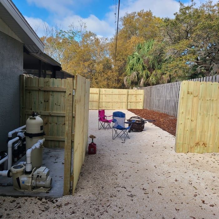 Backyard with wooden fences, gravel ground, pool equipment, fire pit, and chairs.