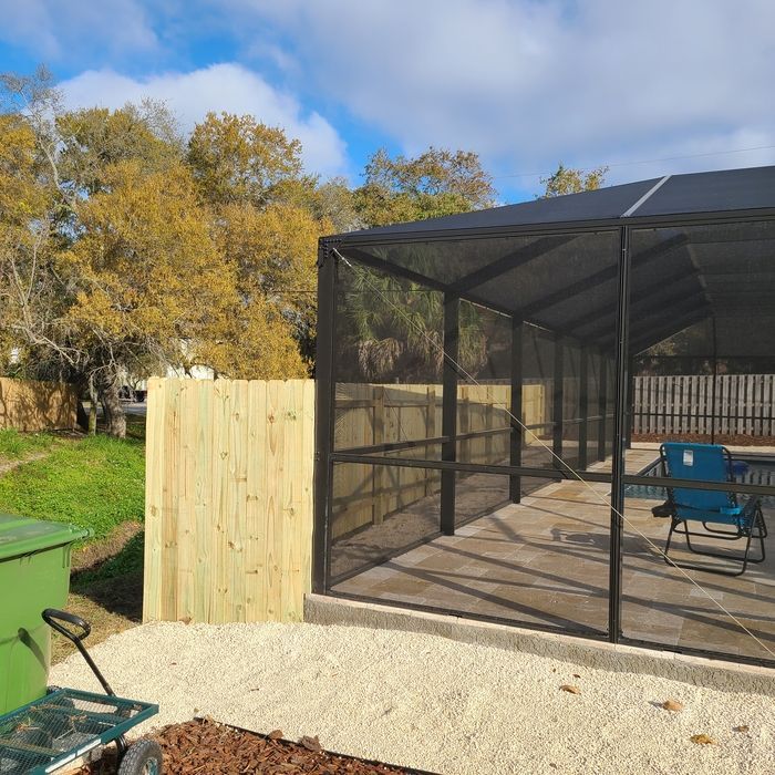 A screened pool enclosure, wooden fence, and yard. Blue sky, trees in the background.