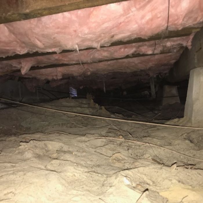 Crawlspace under a house with pink insulation, exposed beams, and dirt floor. Dark and dim interior.