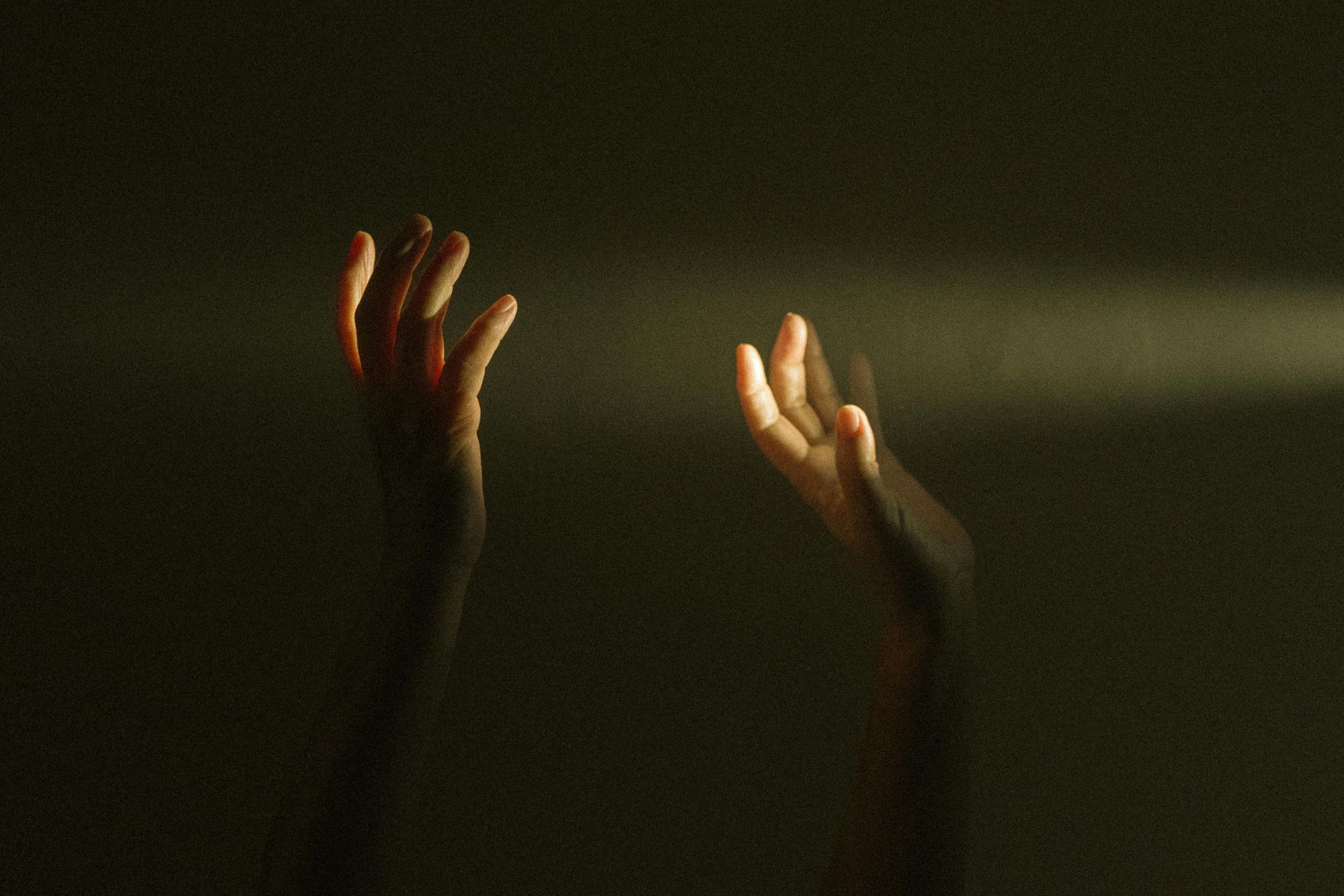Two hands reaching up towards a ray of light, against a dark background.