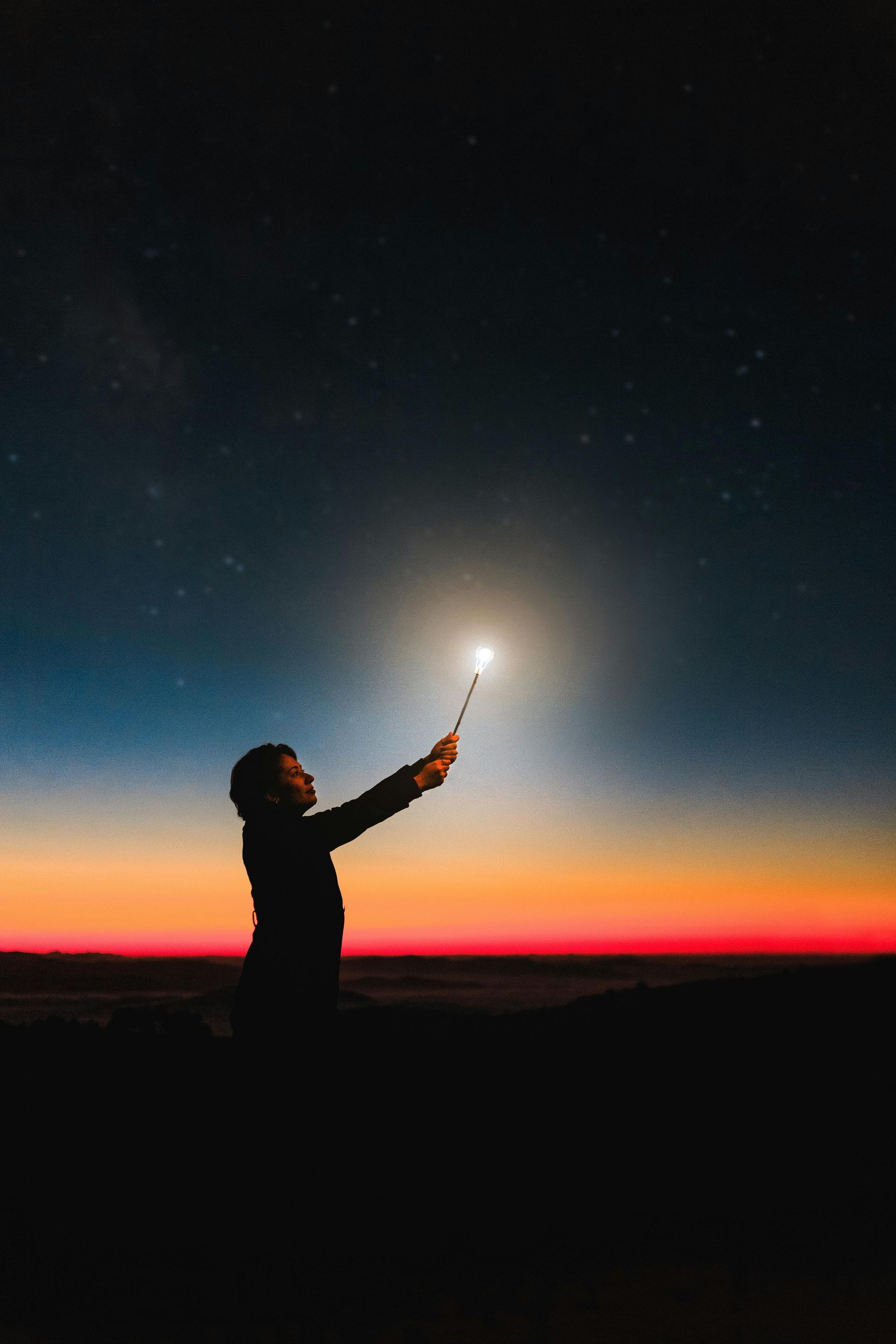 Person holds up wand, glowing in the night sky with a colorful sunset in the background.