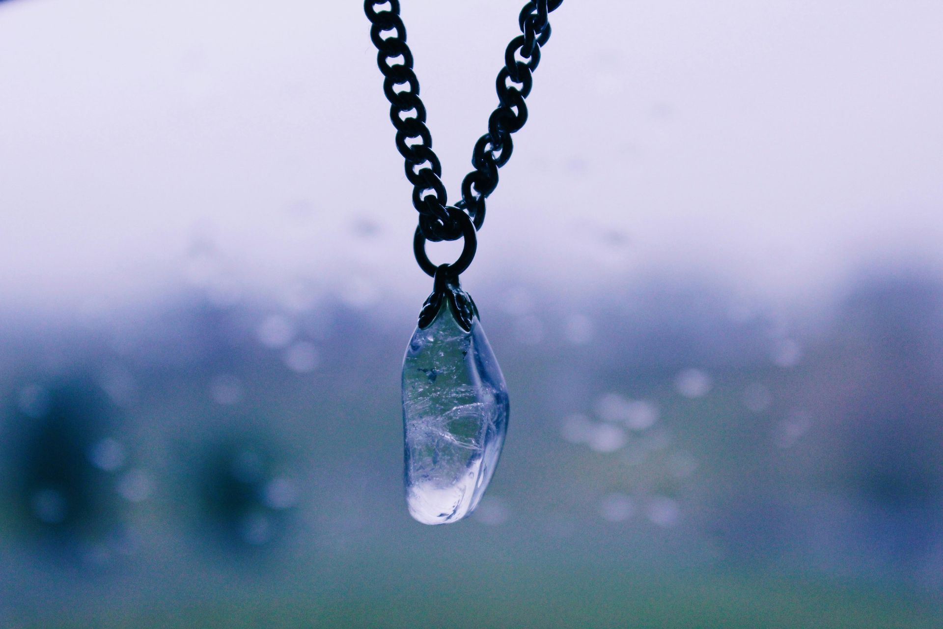 A clear crystal pendant on a black chain hangs in front of a blurry, misty background.