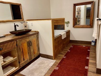 Rustic bathroom with wooden accents. Features a vanity with a dark basin, a built-in tub, and a red rug.
