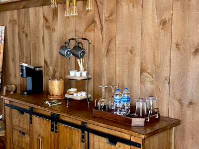 Wooden cabinet with a coffee station set up against a wood-paneled wall. offee maker, mugs, cups, water bottles, and glasses.