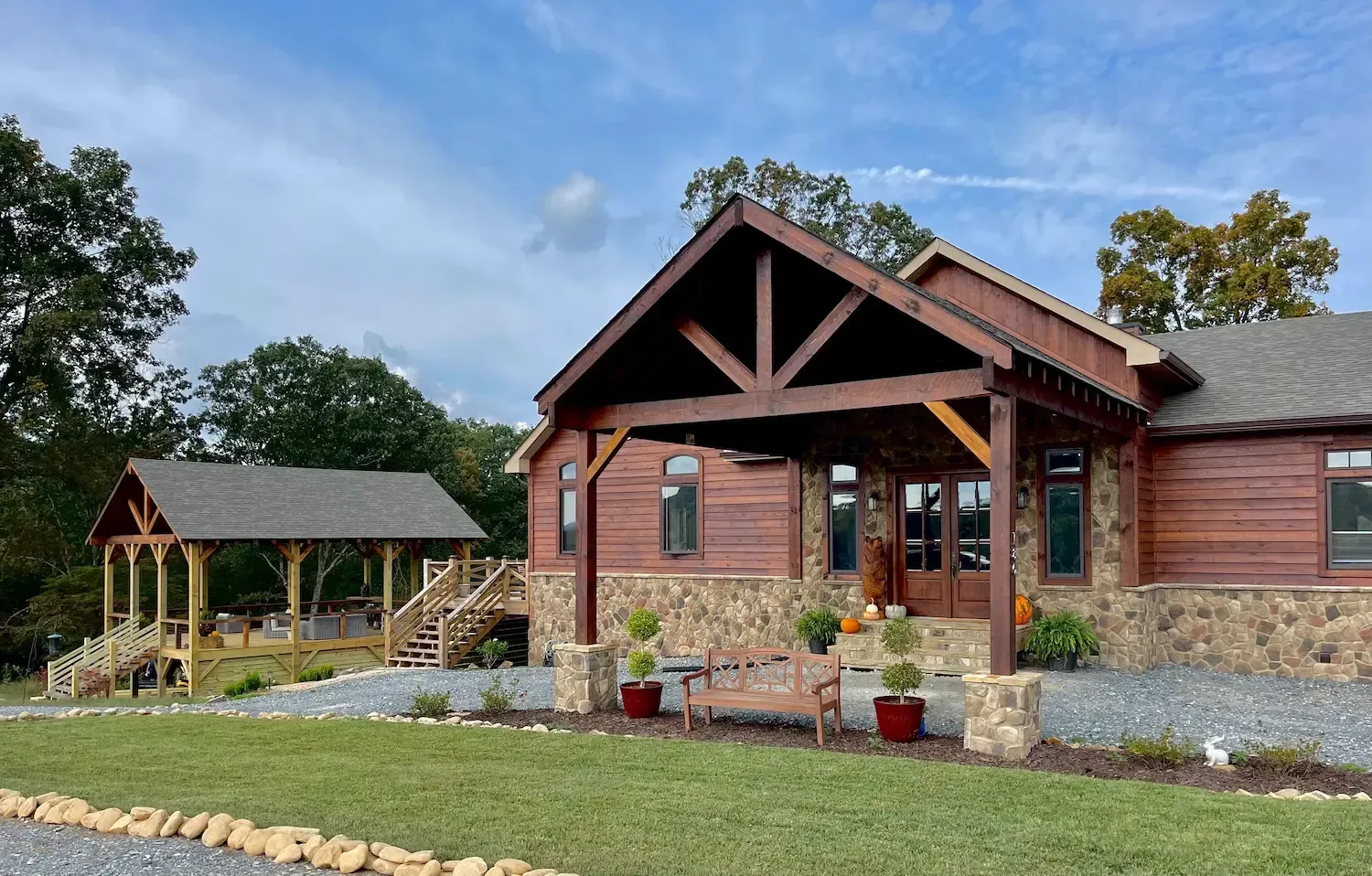 A wooden lodge with a stone facade, porch, and gazebo set on a grassy lawn under a blue sky.