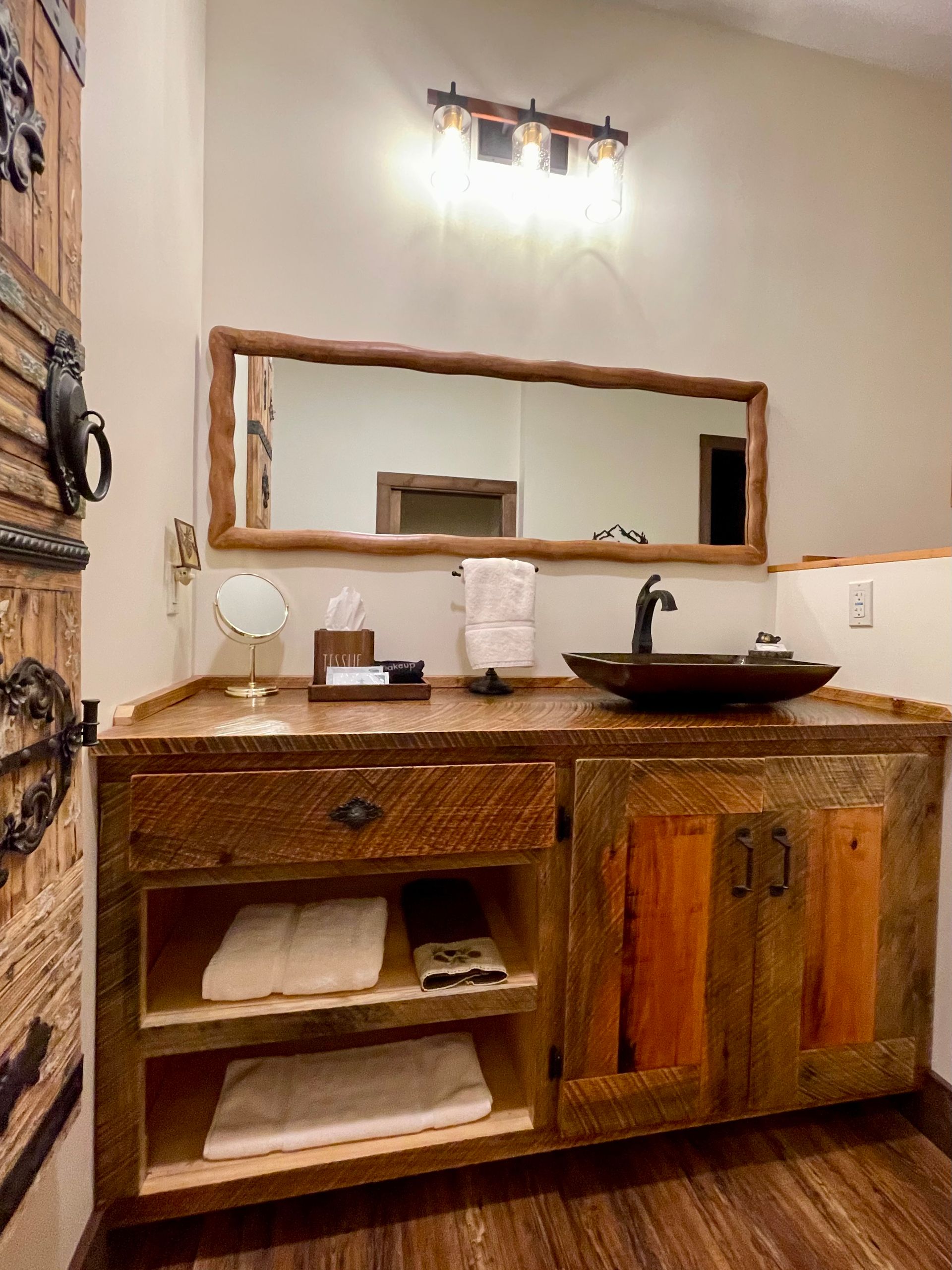 Rustic bathroom with wooden vanity, vessel sink, and a large mirror. Beige walls and brown floors.
