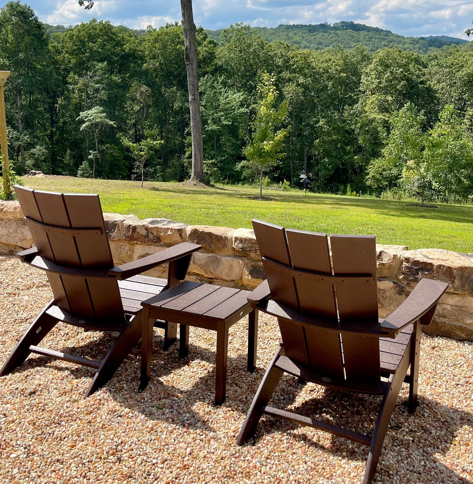 Two brown Adirondack chairs and a small table sit on a stone patio overlooking a green lawn and forest.
