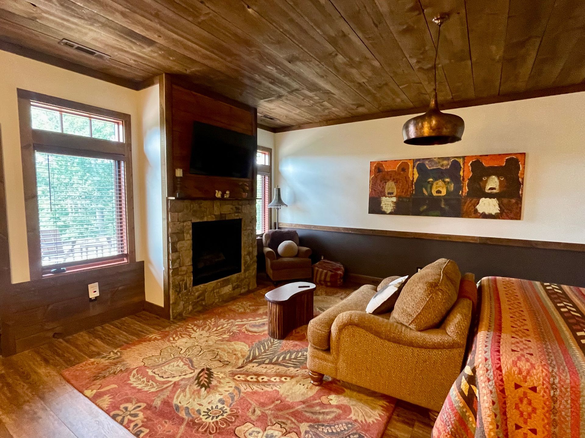 Cozy cabin bedroom with fireplace, TV, and seating area featuring a red patterned rug and artwork of bears. Wooden ceiling