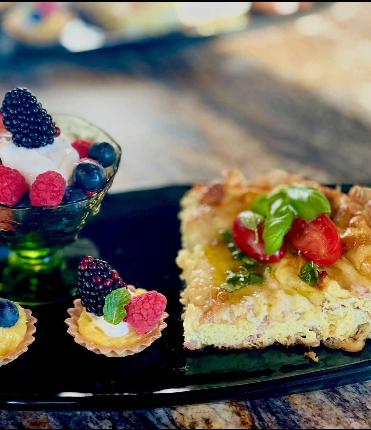 Black plate with a quiche, a glass of berries and whipped cream, and berry tarts on a rustic wooden surface.