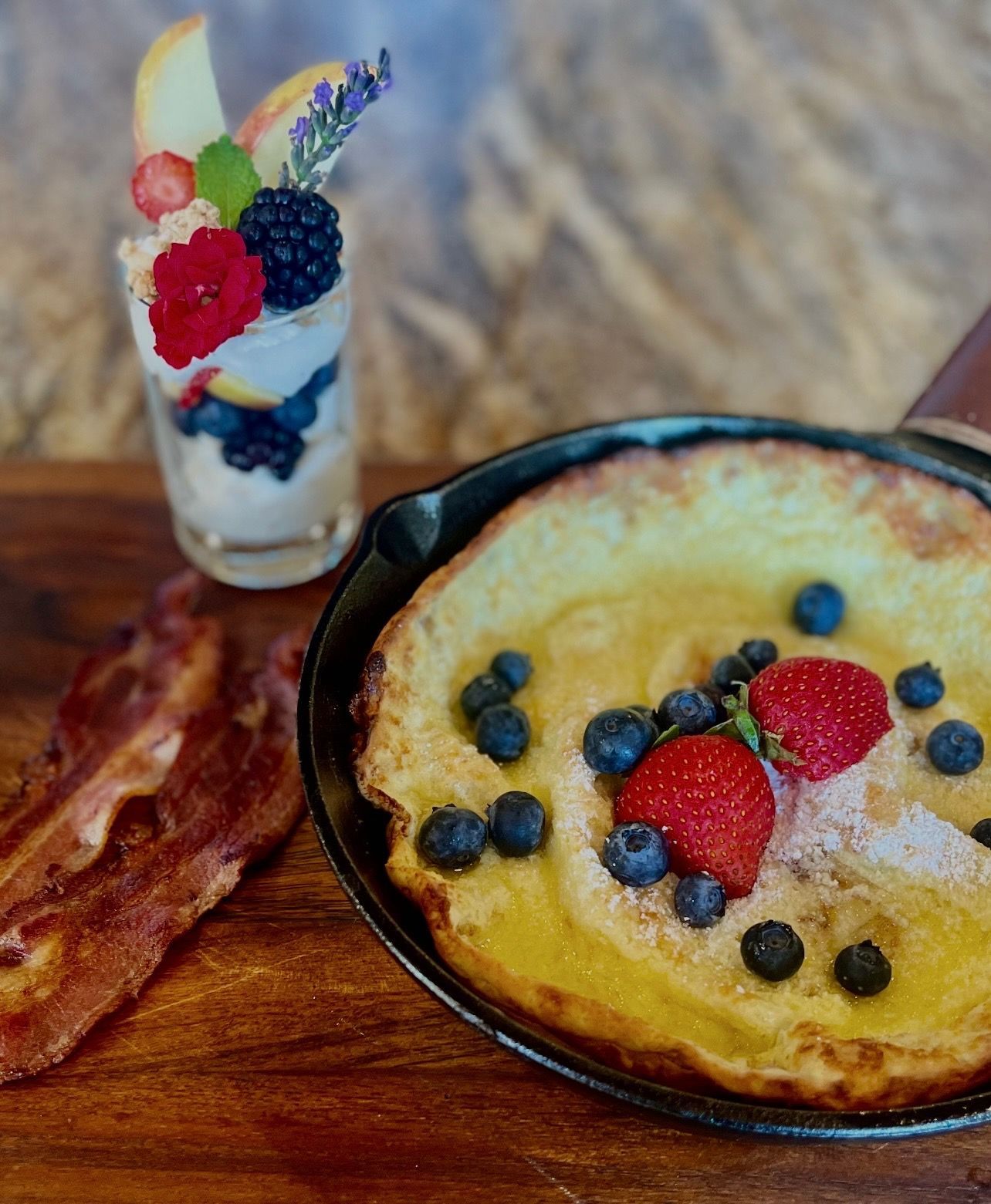 Dutch baby pancake topped with blueberries and strawberries in a cast iron skillet, with a yogurt parfait and bacon.