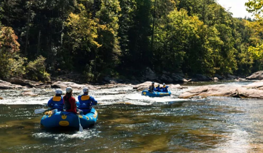 Three people in a blue raft paddle down a river, with another raft in the distance; green trees line the banks