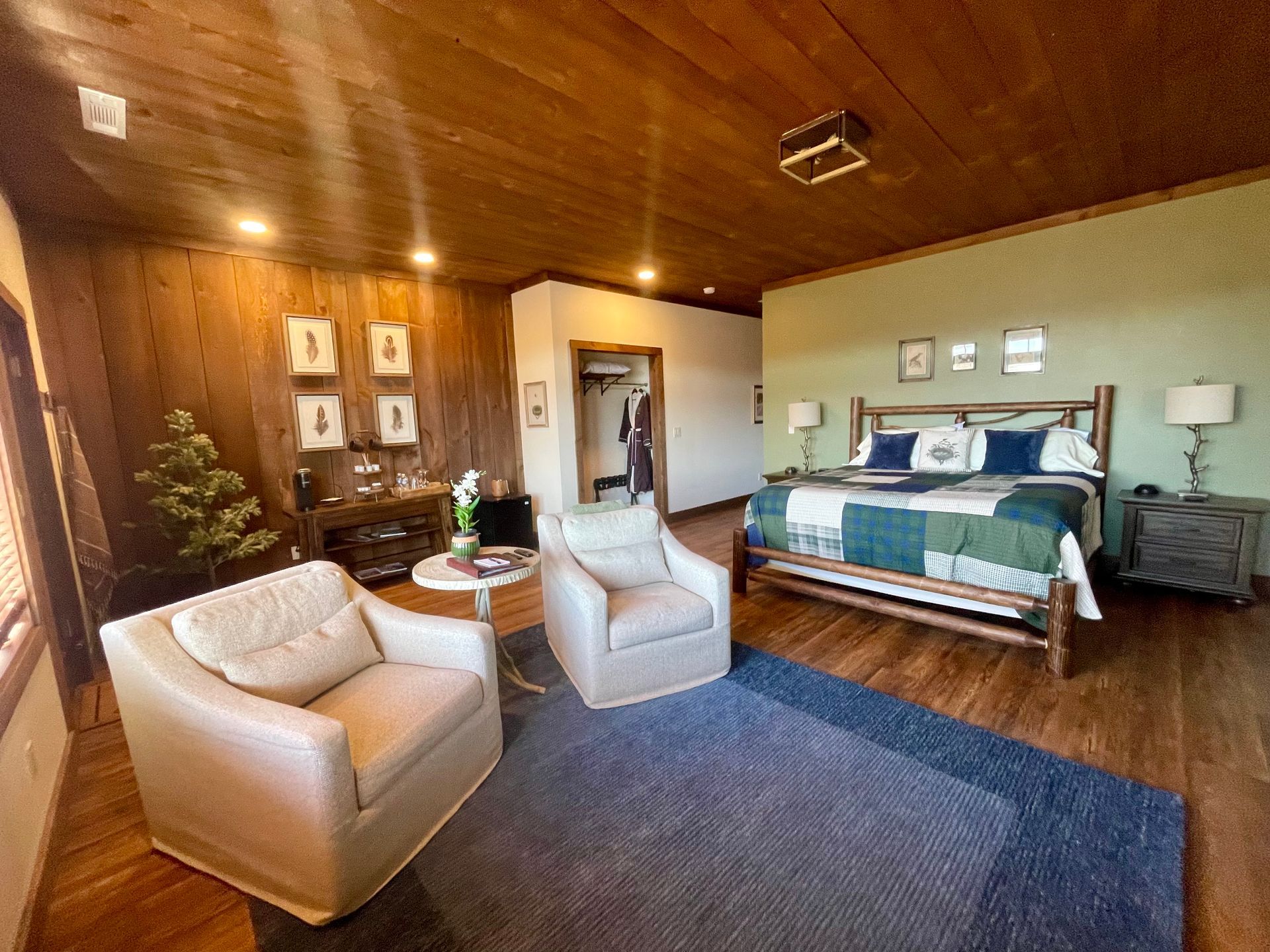 A spacious bedroom with wood paneling, two white armchairs, a bed with a patchwork quilt, and a closet.