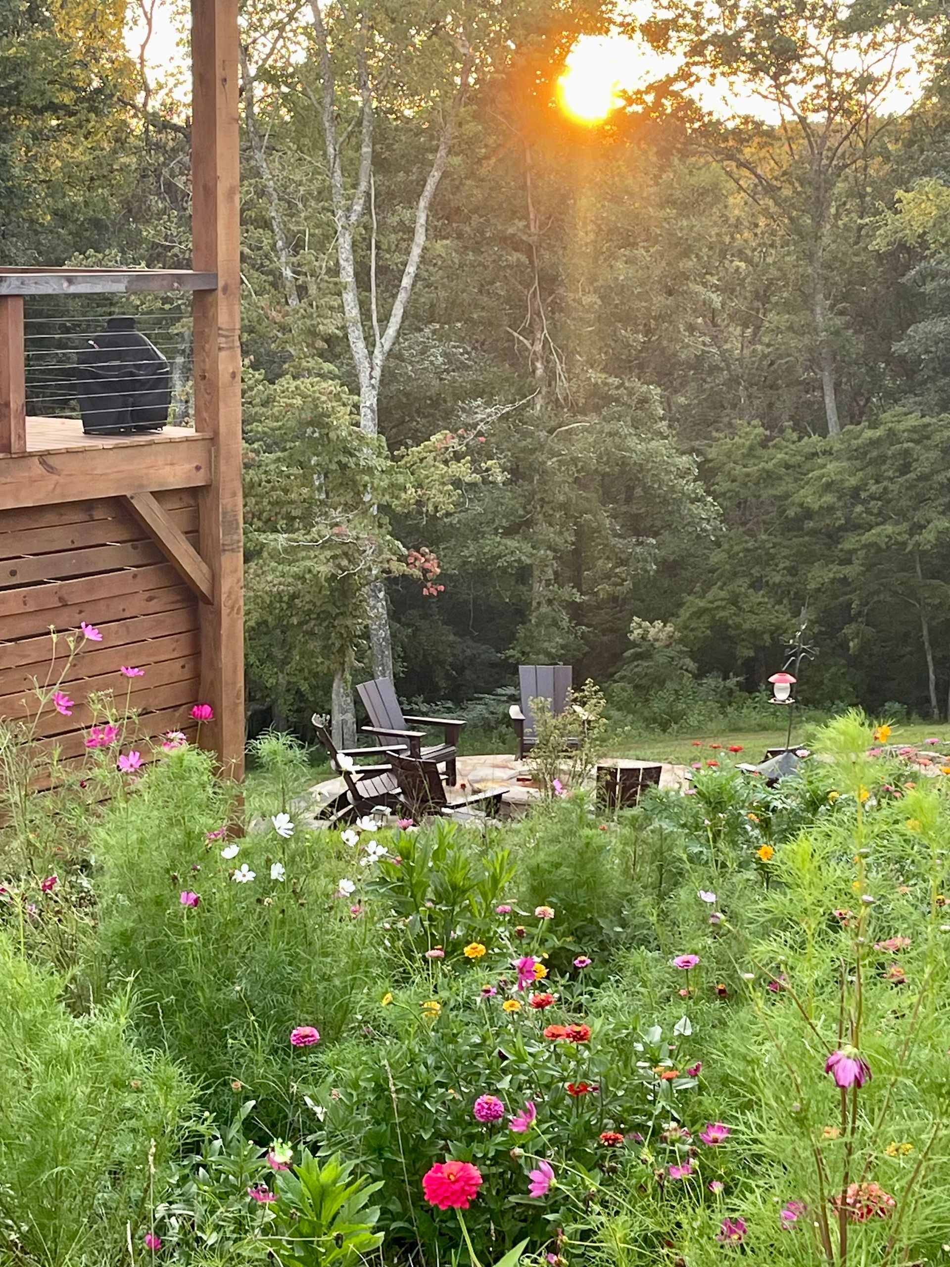 A lush flower garden with a wooden deck and chair set. The sun shines through trees.