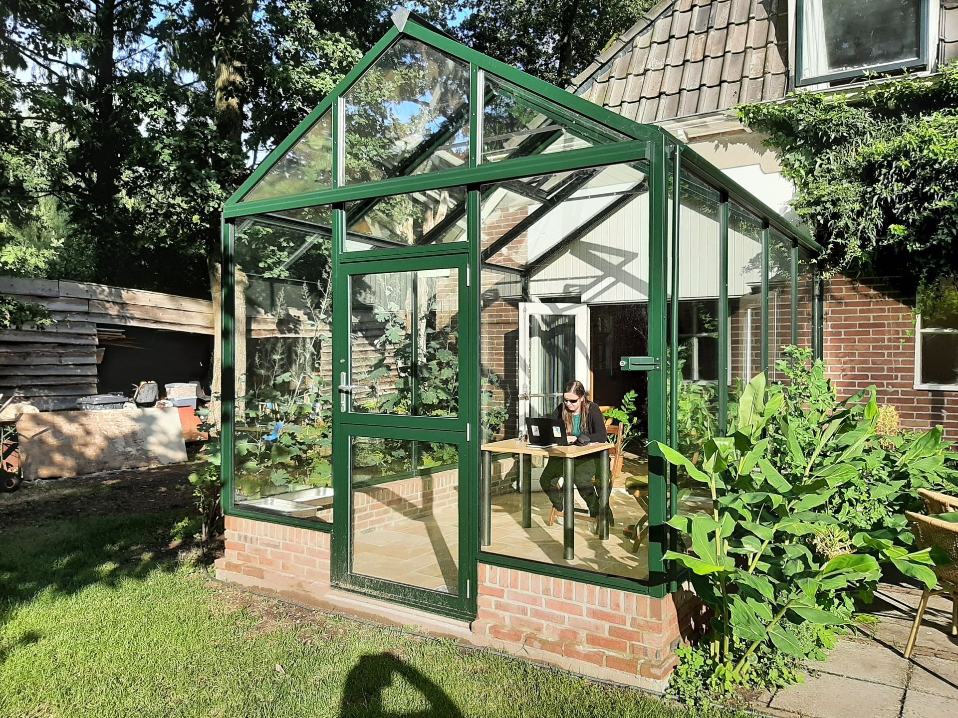 Kas met glazen wanden en deur, vastgemaakt aan een bakstenen huis, planten aan de voorkant.