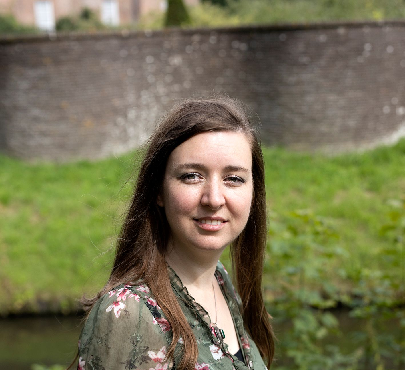Een vrouw met lang bruin haar lacht, terwijl ze buiten staat bij het water en een bakstenen muur.