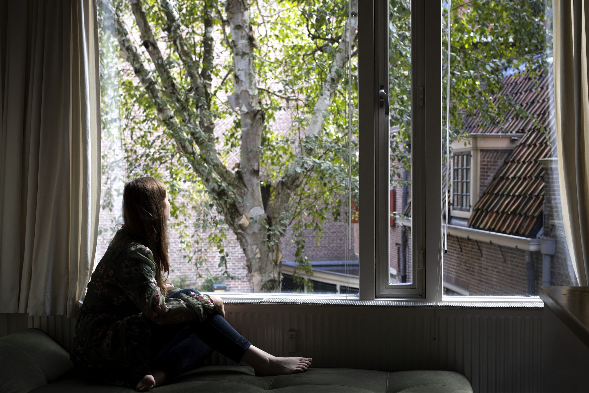 Een vrouw zit bij het raam en kijkt naar buiten, naar een boom en gebouwen. Het zonlicht stroomt naar binnen.