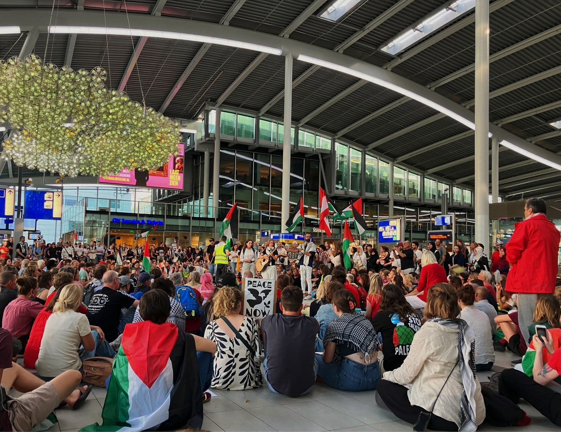 Protest op een treinstation; menigte zit op de grond en zwaait met Palestijnse vlaggen.