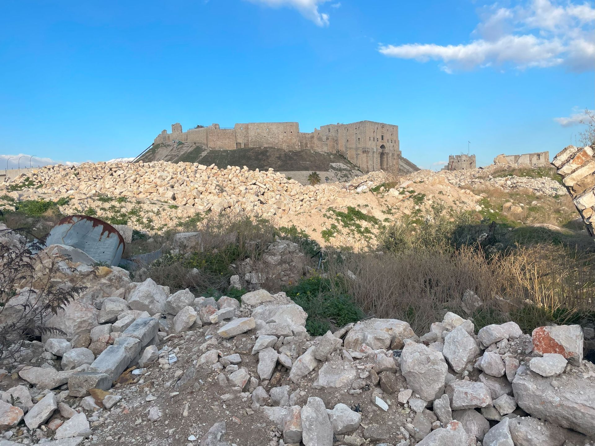 Verwoeste gebouwen en puin op de voorgrond: een kasteel op een heuveltop onder een blauwe lucht.