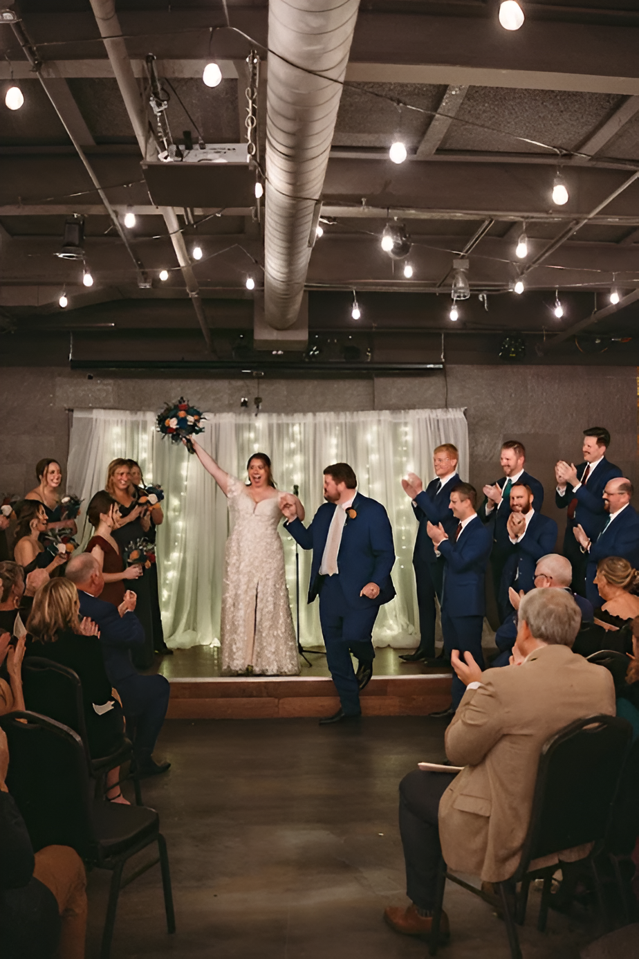 Bride and groom celebrate, walking down an aisle as guests applaud; reception hall with string lights.