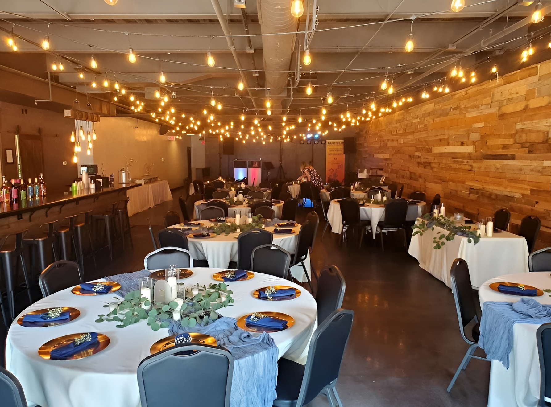 Event space with tables set for a gathering, strung lights overhead. Tables have gold chargers, blue accents, and centerpieces.