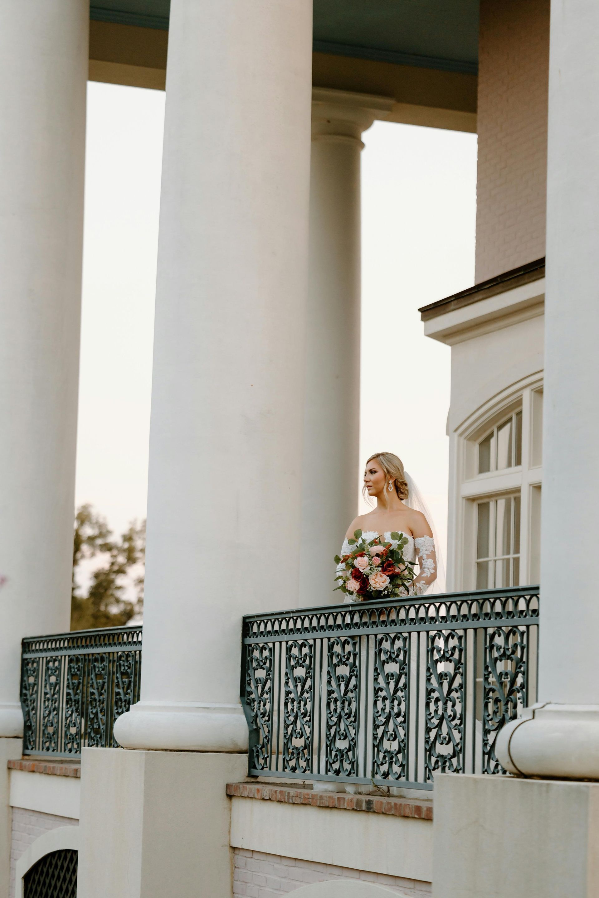 Bride holding bouquet on a balcony, surrounded by white columns.