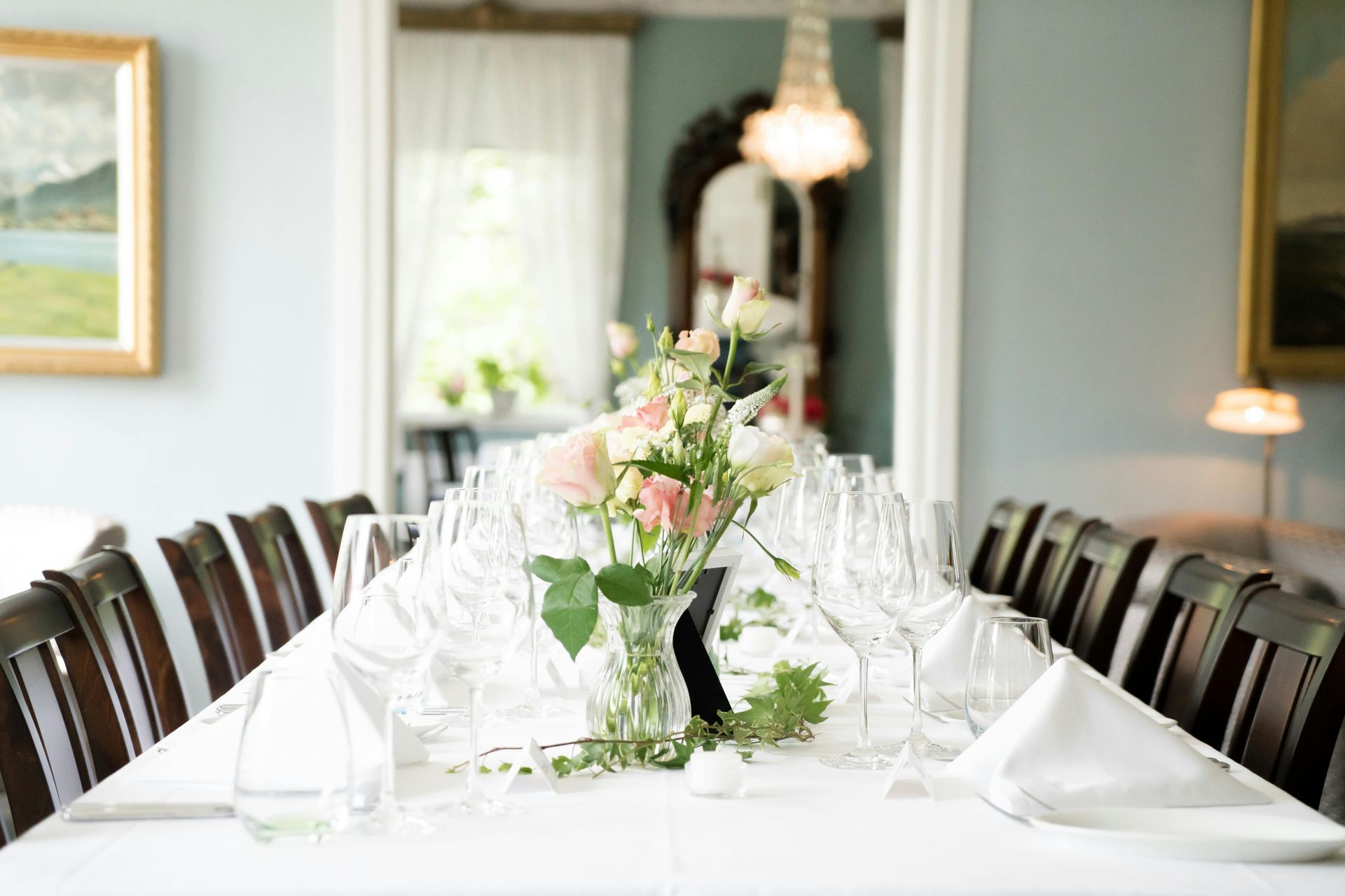 Elegant table set for a meal, with a floral centerpiece. Chairs, glasses, and napkins line the white tablecloth in a room.