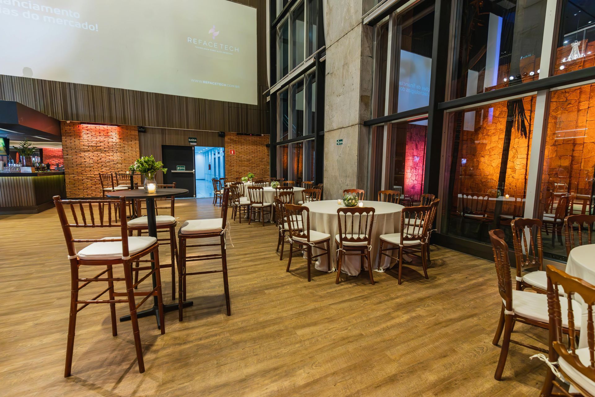 Event space with tables, chairs, and large windows. Wooden floors, brick walls, and ambient lighting.