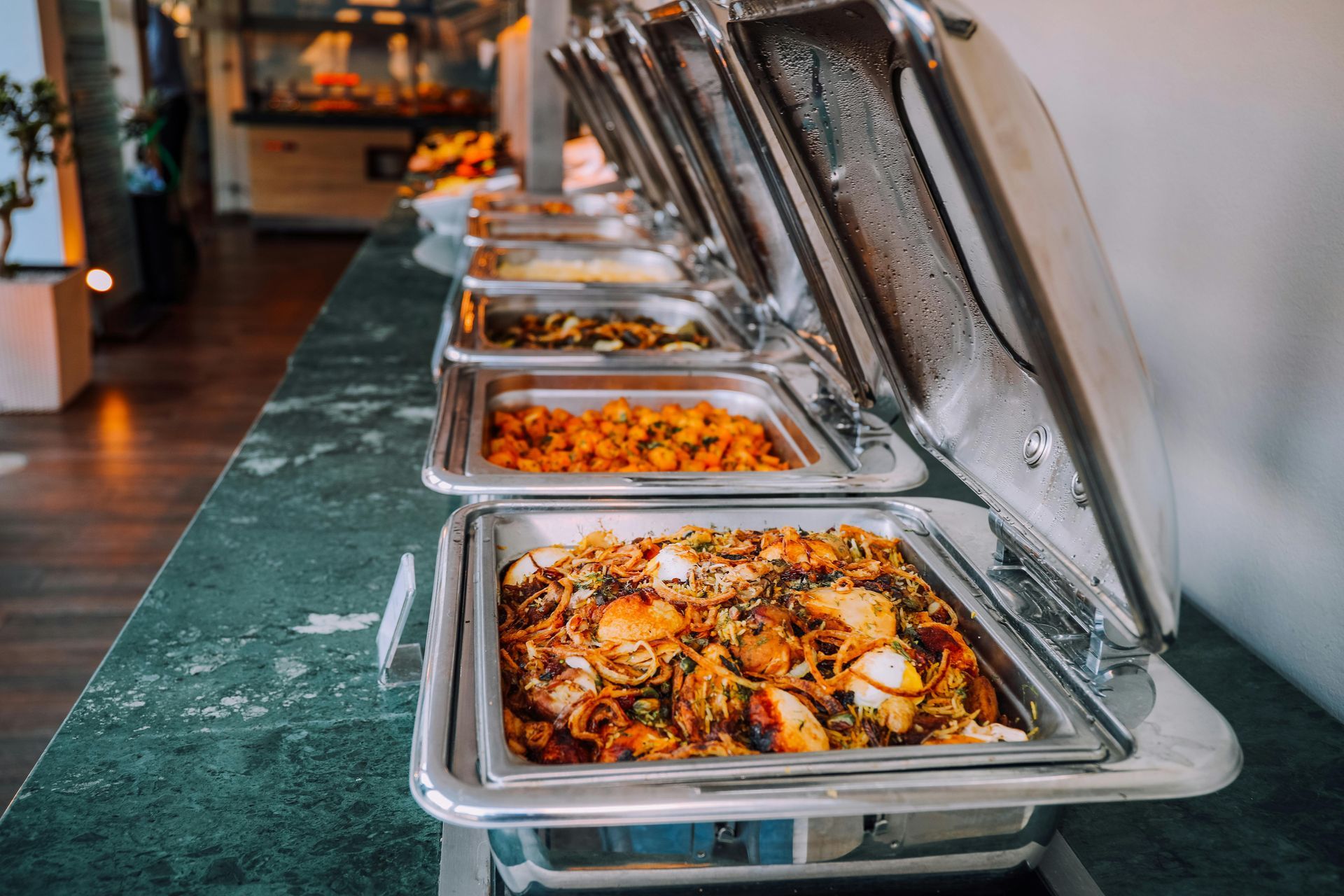 Buffet with various dishes in silver serving trays on a green countertop.