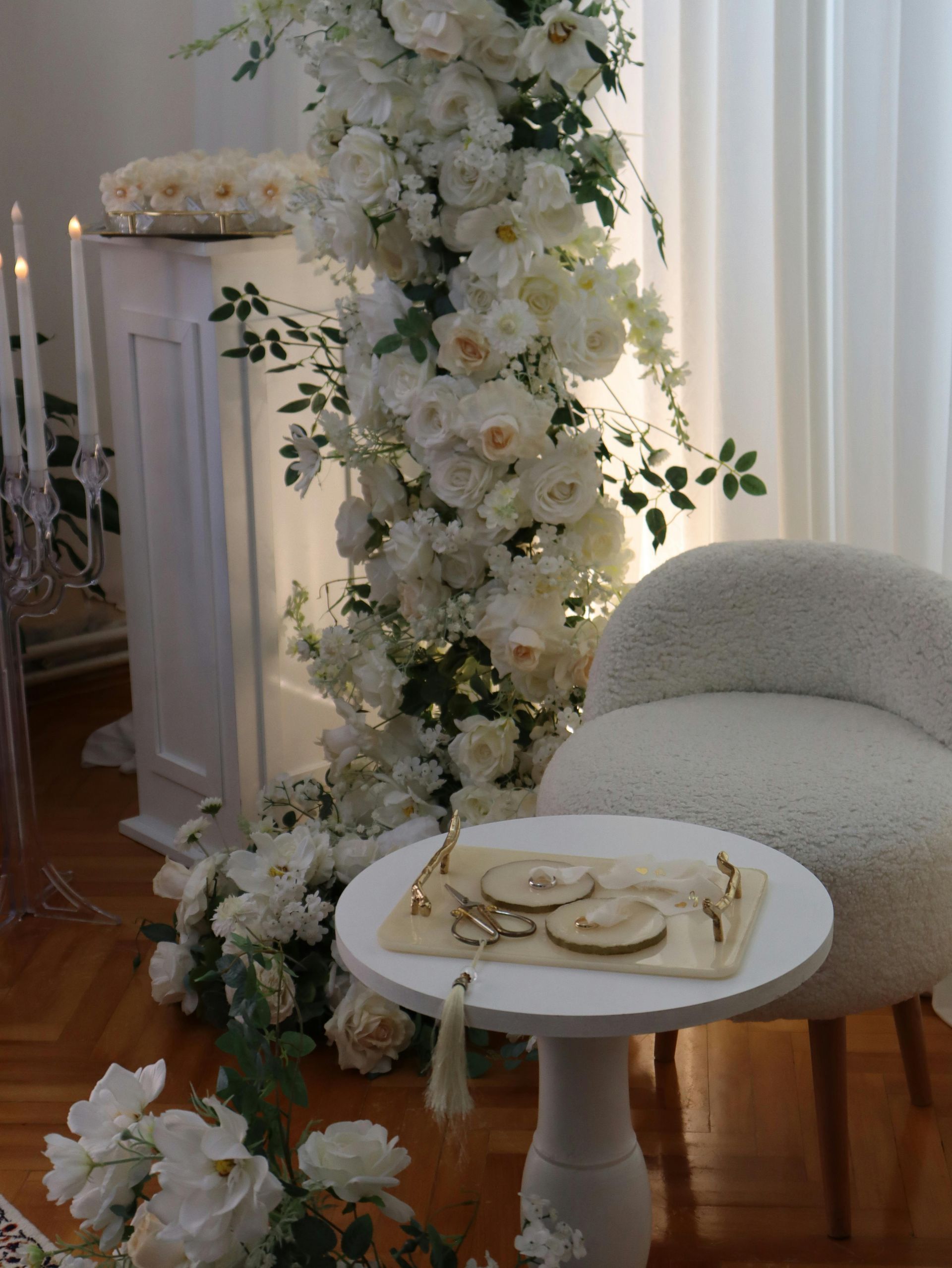 A white floral arch next to a small table with rings, a fluffy chair, and candles in an elegant setting.