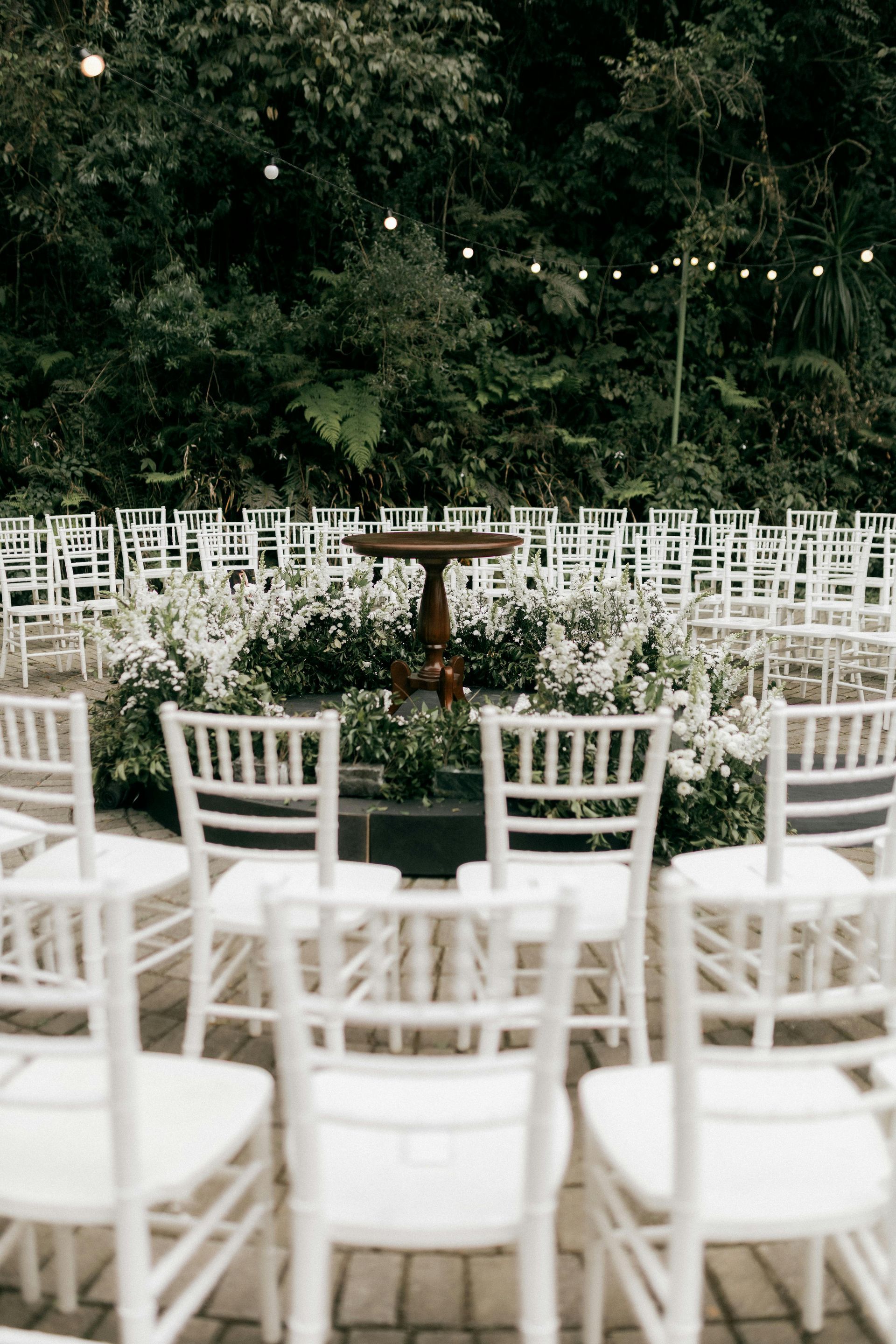 Wedding ceremony setup: White chairs face a floral altar with a dark table. Lush green backdrop.