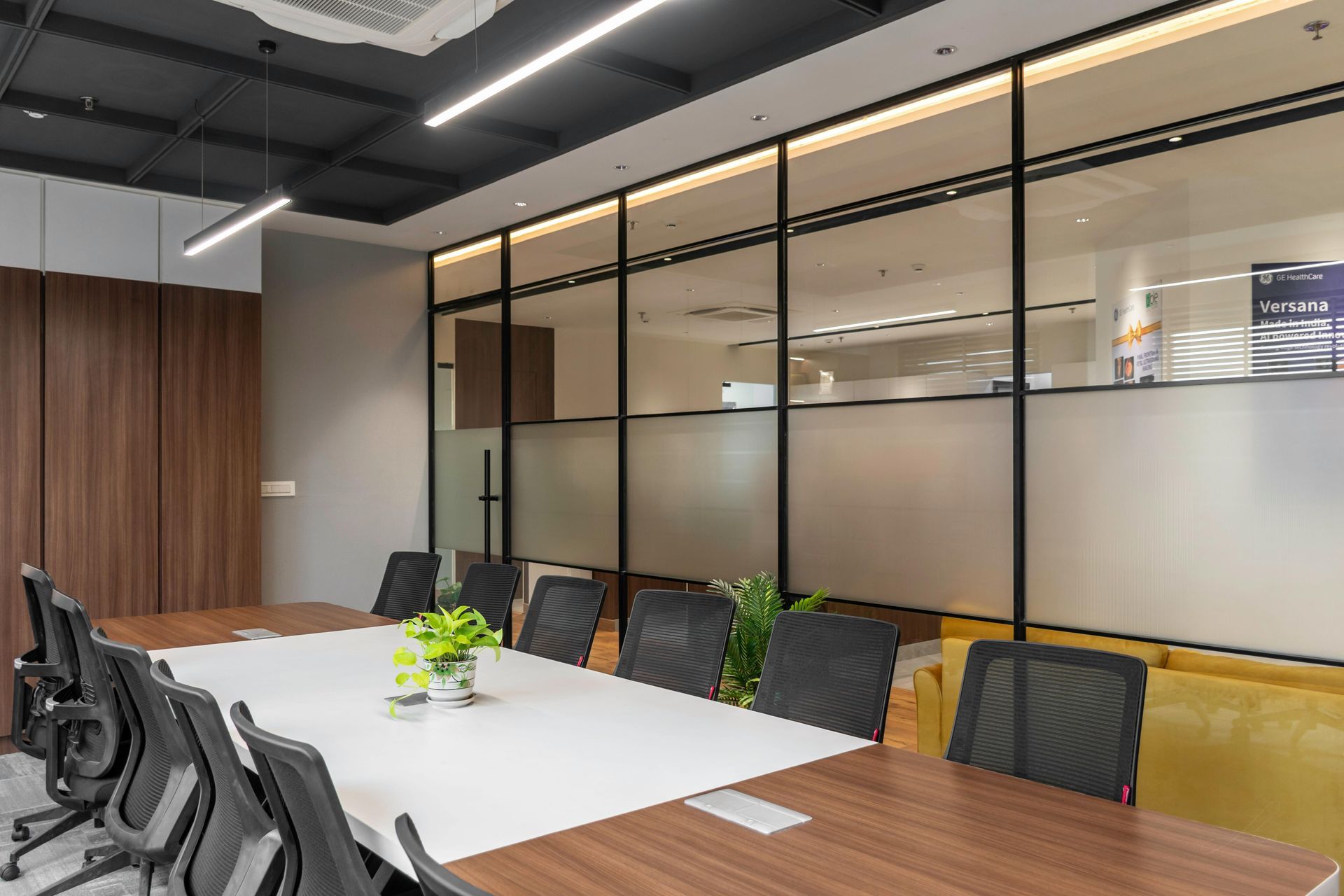 Conference room with long wooden table, black chairs, frosted glass wall, and modern lighting.