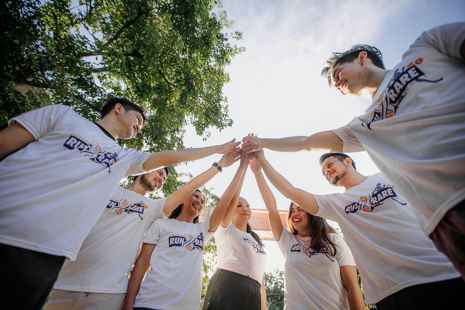Group of people in white shirts, hands together in a circle. Outdoors, under a tree, sunny.
