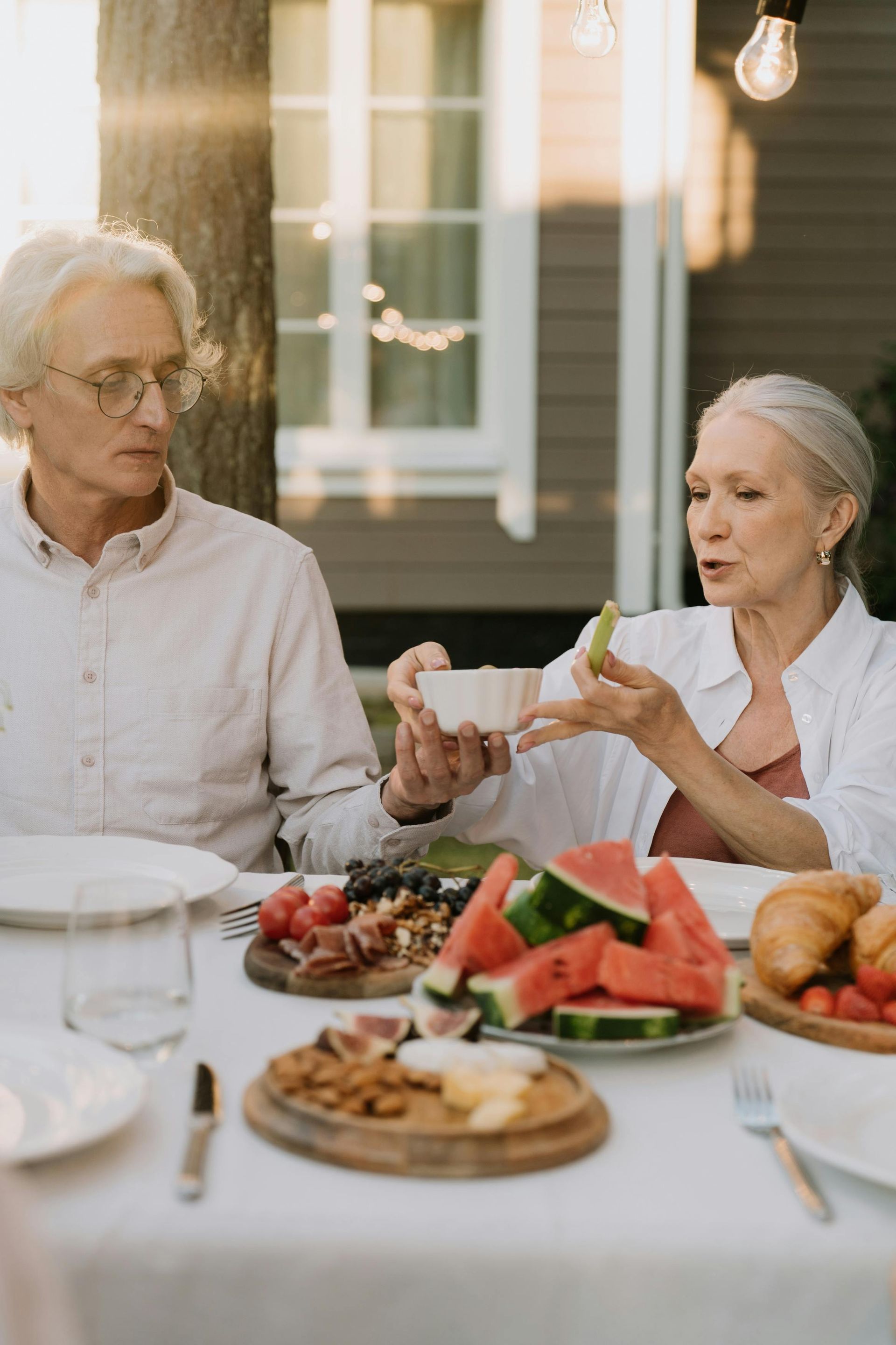 Elderly couple at an outdoor table, eating a meal with fruit. Woman gestures while speaking.