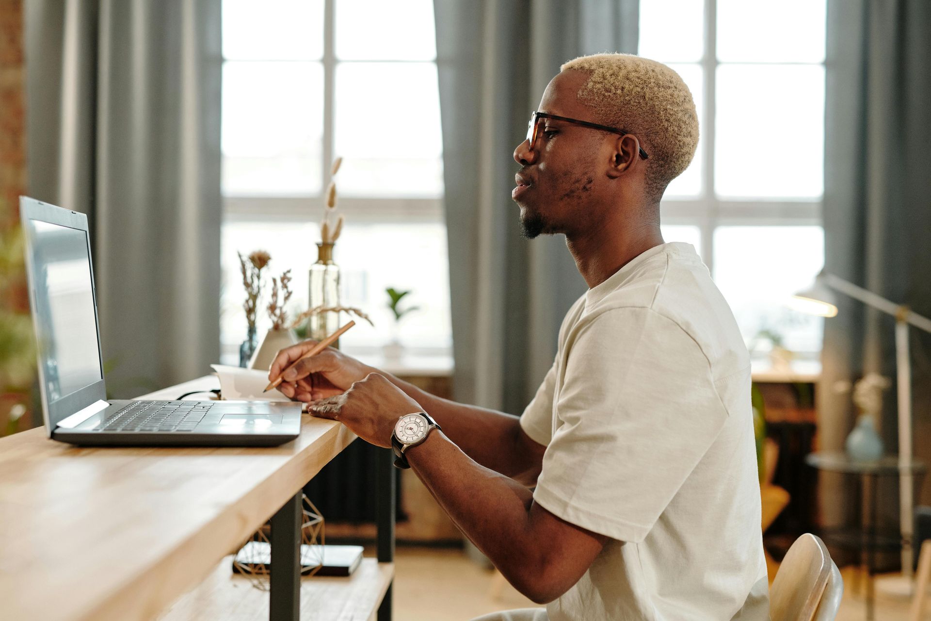 Man with blonde hair working on a laptop at a wooden desk, writing in a notebook. Interior shot.