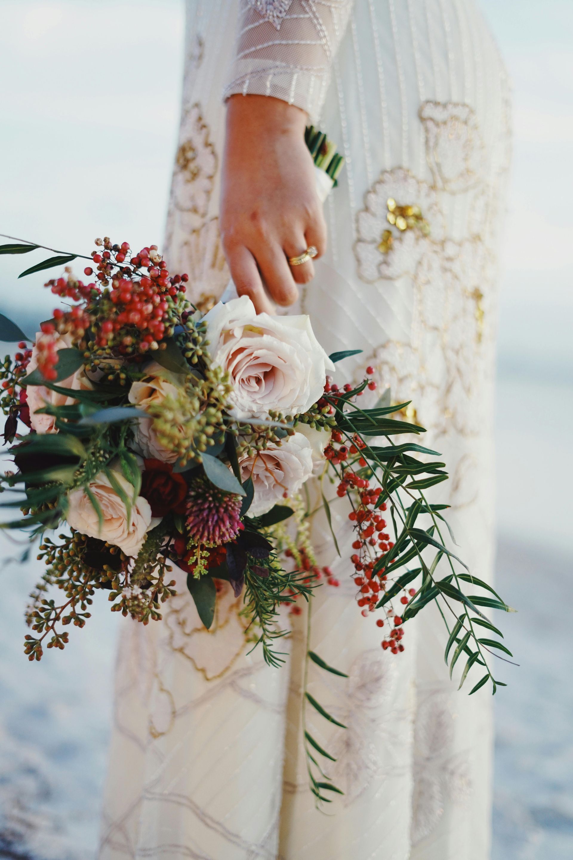 Bride holding a bouquet of roses and berries, wearing a white beaded dress on the beach.