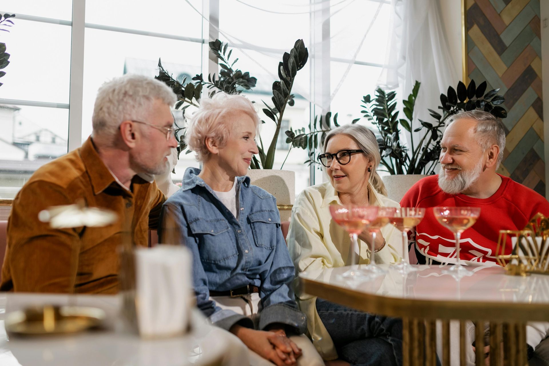 Four older adults at a table, some with pink drinks, talking. Interior, with plants and neutral tones.